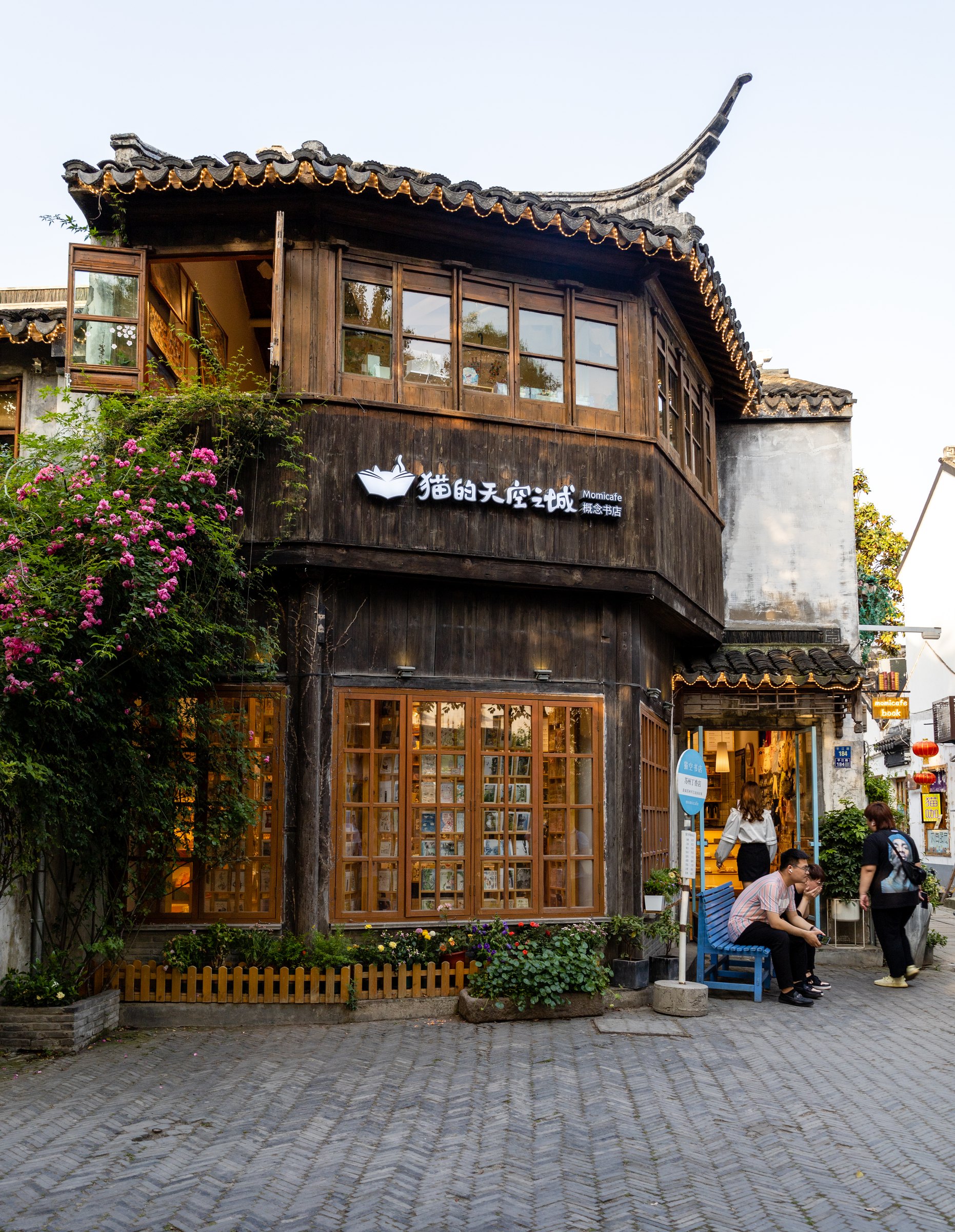 街角两层木质店铺有翘起的灰瓦屋檐,粉色花枝沿左侧攀生,门边有人停留。