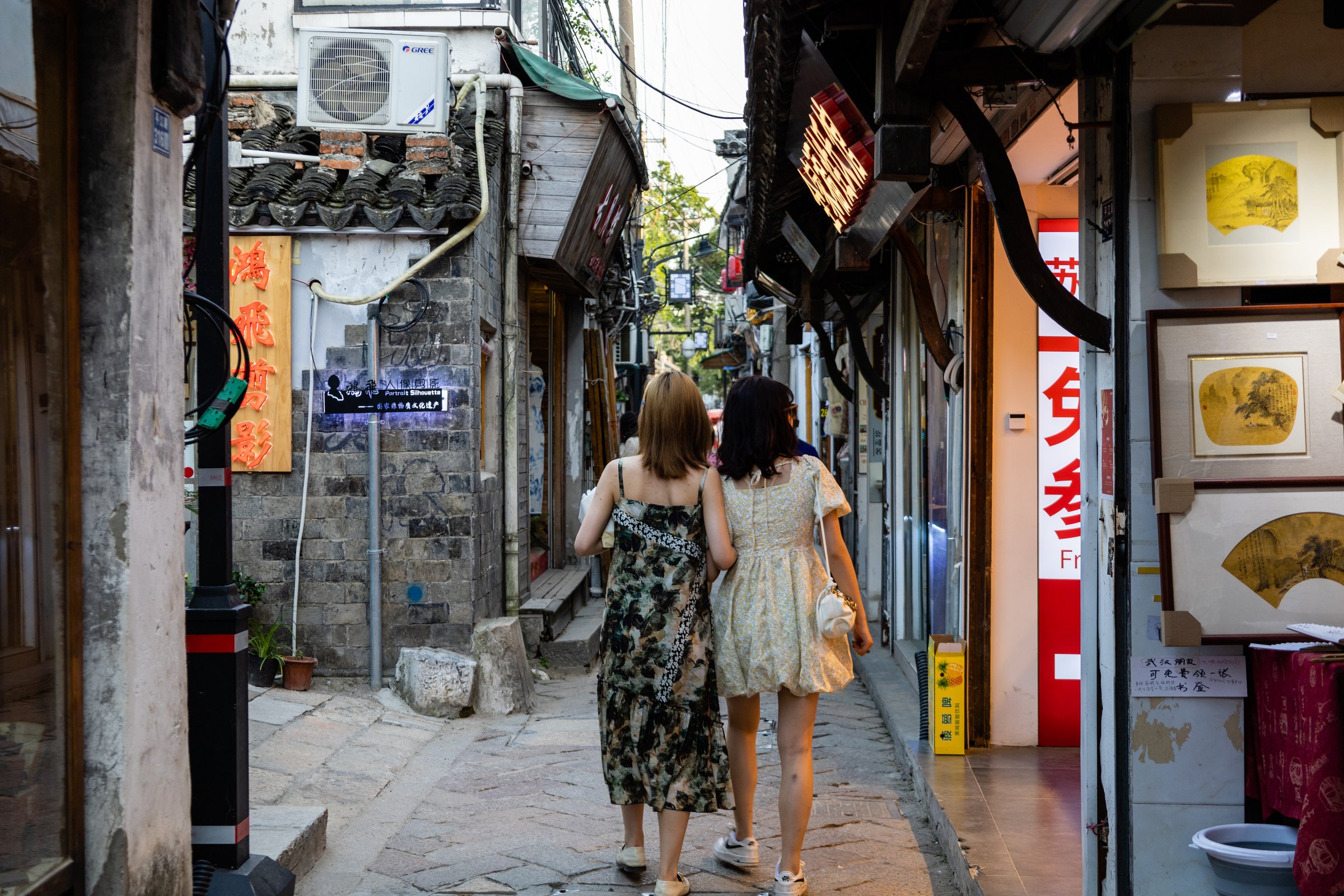 两名穿花纹连衣裙的人背向镜头走进窄巷,右侧店面展示装框扇面。