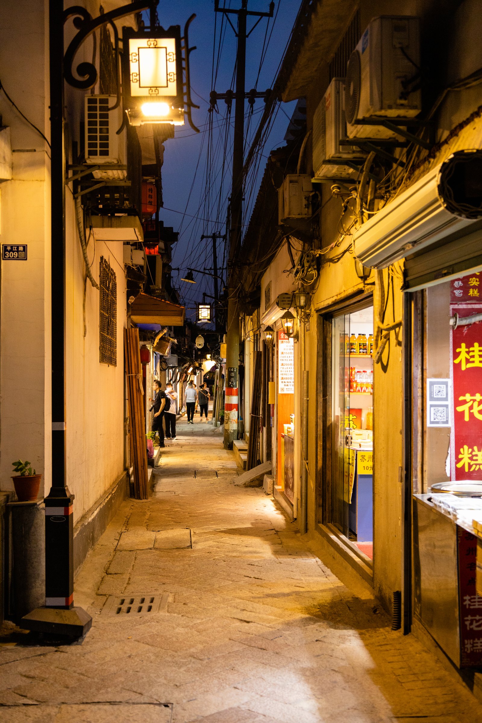 深蓝天空下的窄巷布满暖色店灯,石板路上有行人,两侧可见空调外机与电线。