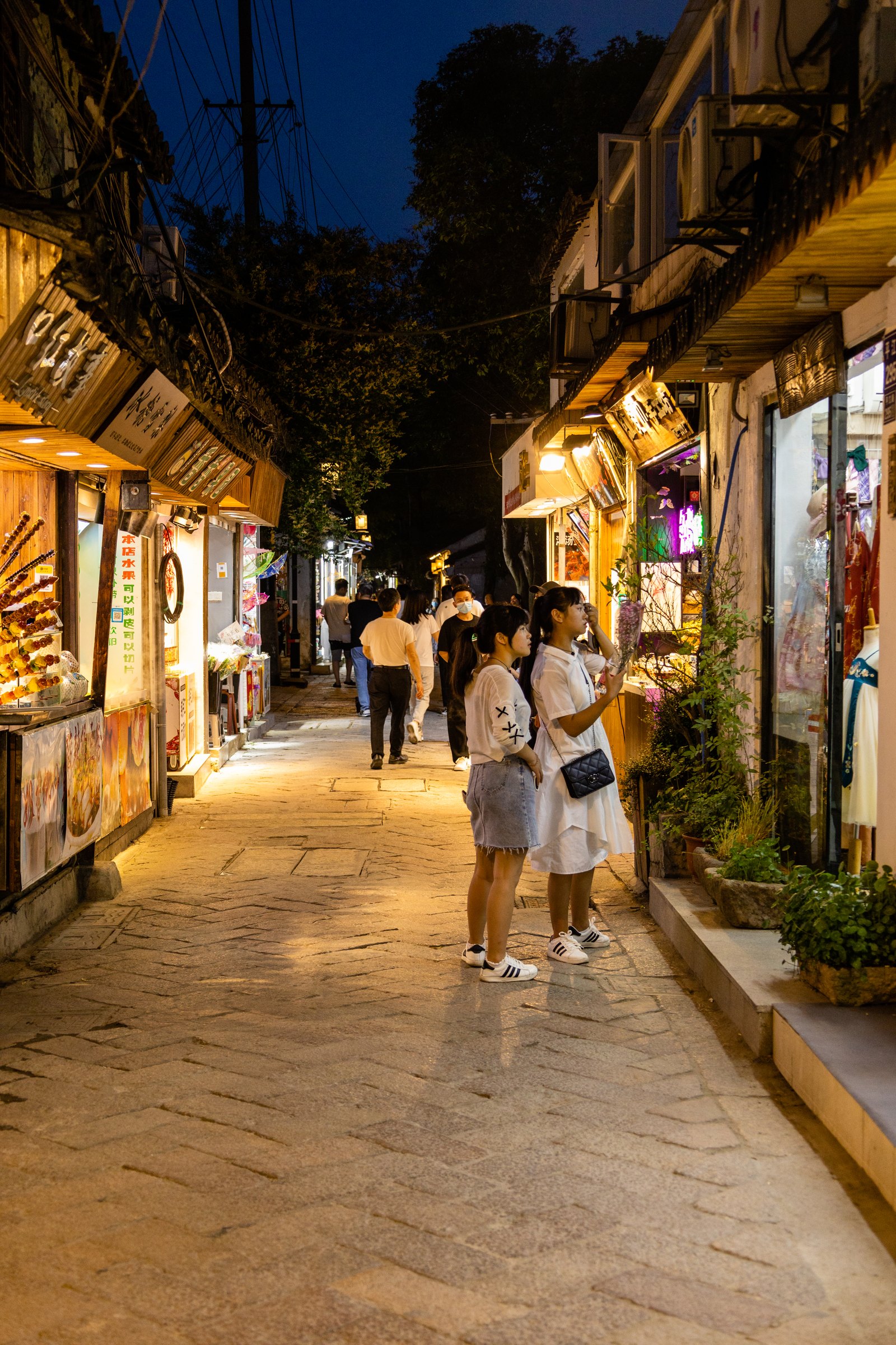 灯光照亮石铺商业小巷,两名人物停在右侧店窗前,远处还有行人。