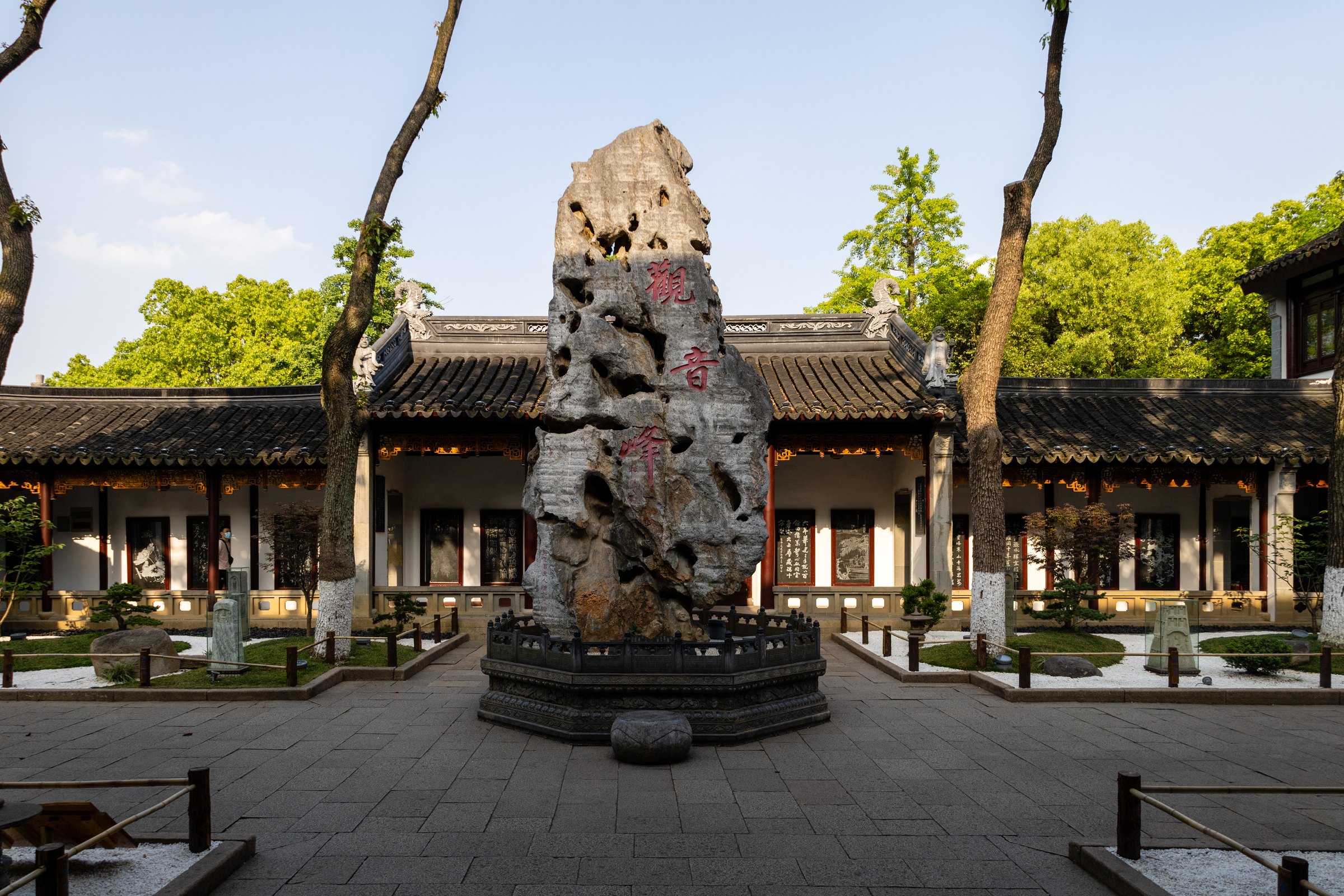庭院中央矗立一块带孔洞的高大石景,后方是灰瓦白墙建筑和树木。