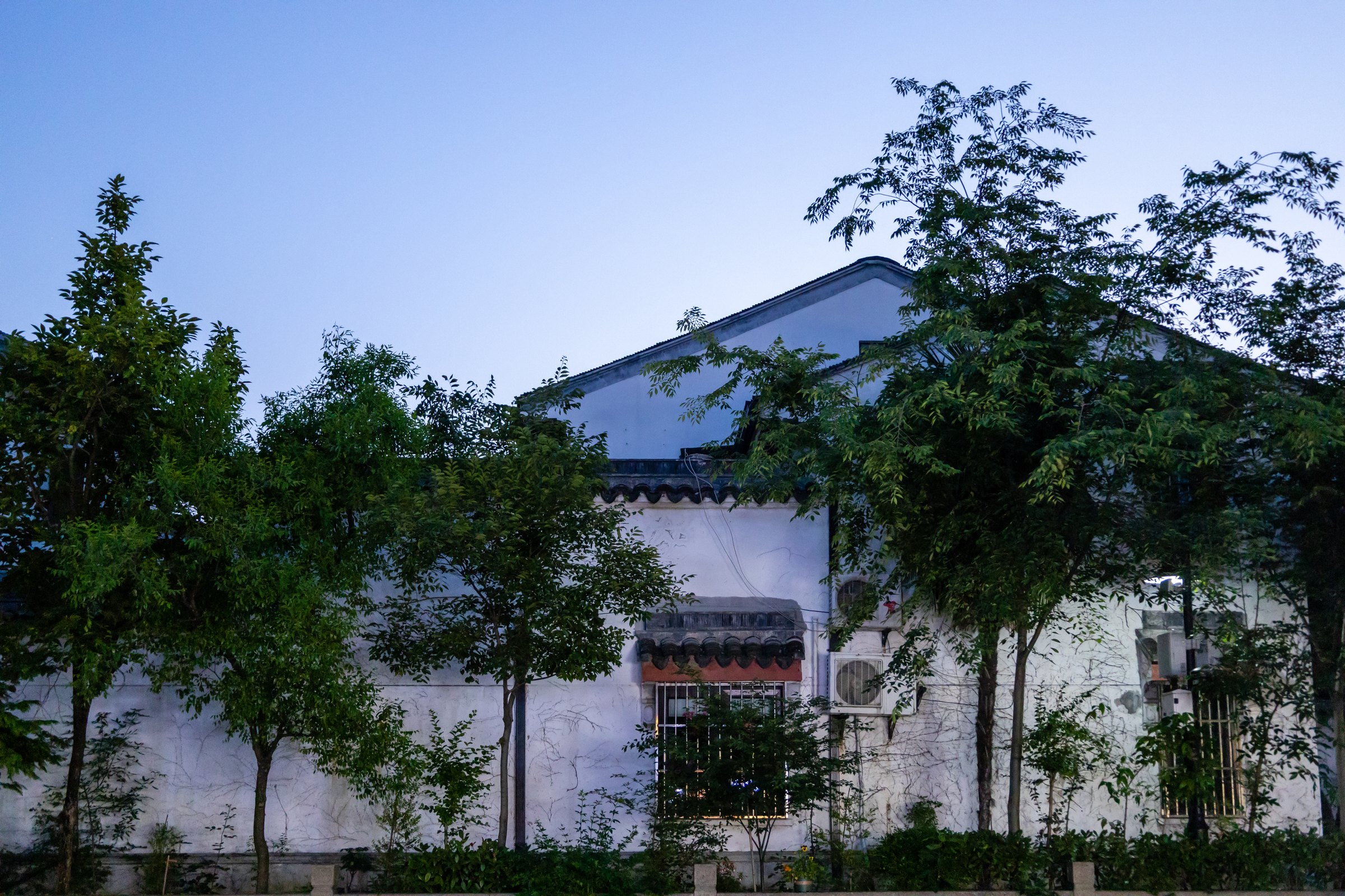 蓝色天空下,几株绿树遮住白墙灰瓦房屋的一部分,墙上可见窗户和空调外机。