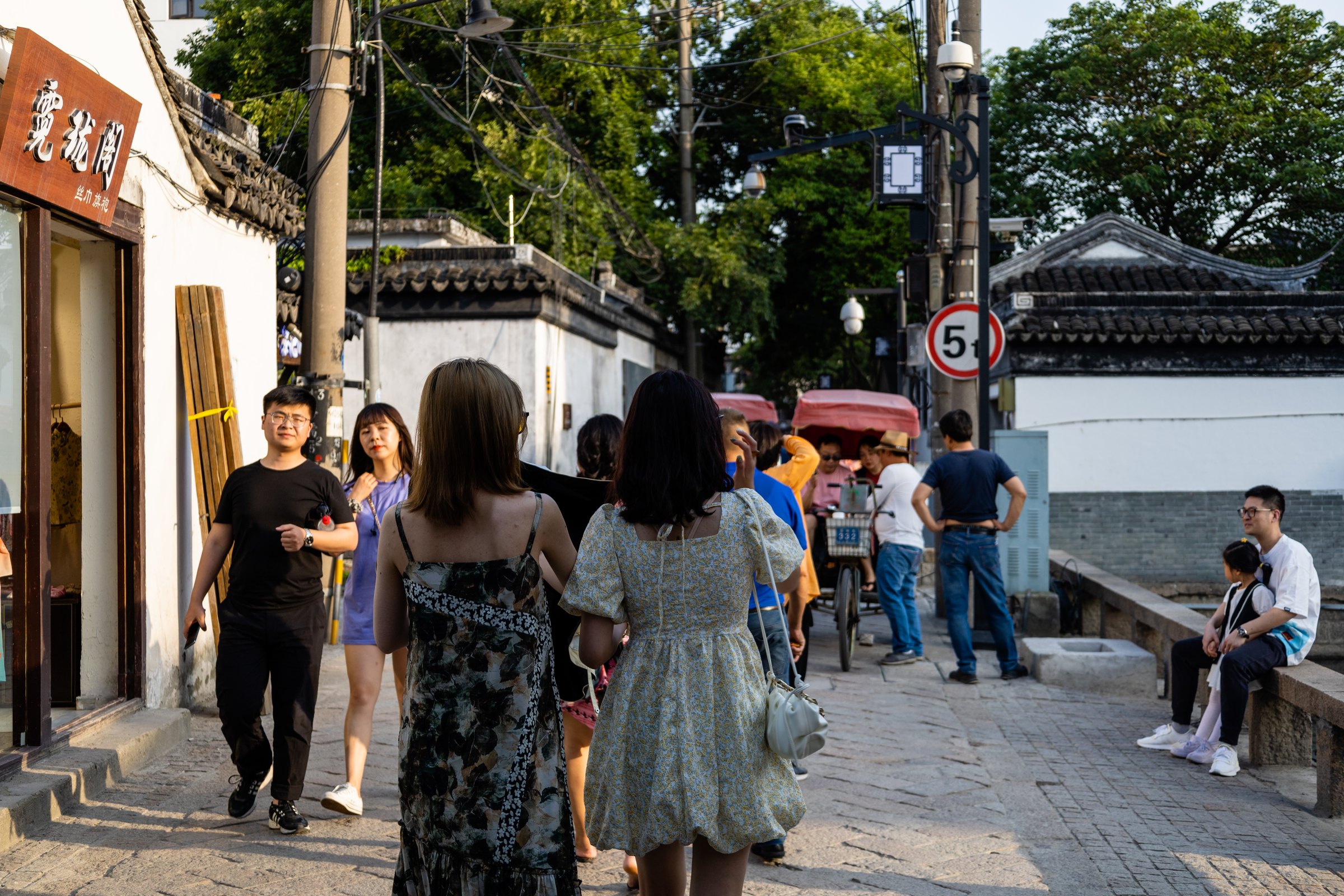 多人行走在白墙灰瓦之间的石铺街道,右侧岸边栏杆旁有人坐着。