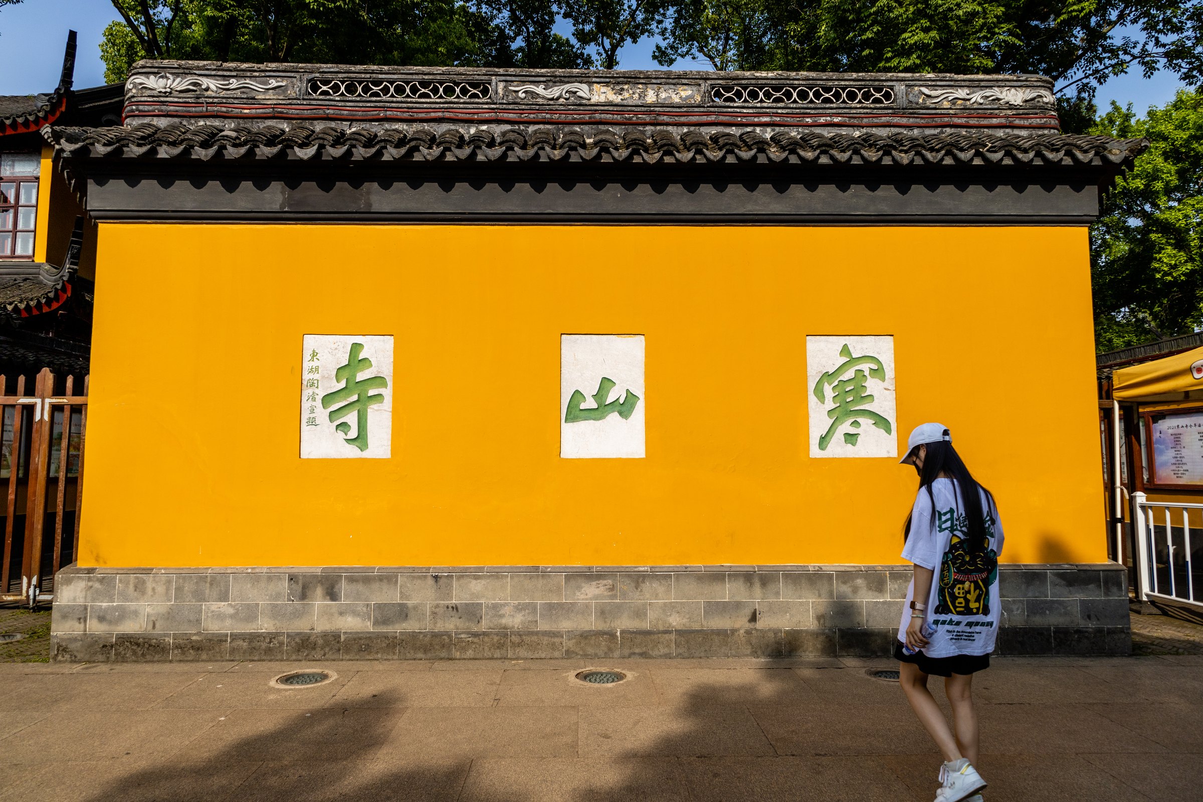 一人戴白色帽子从明黄色墙前经过,墙上嵌有三个绿色大字。