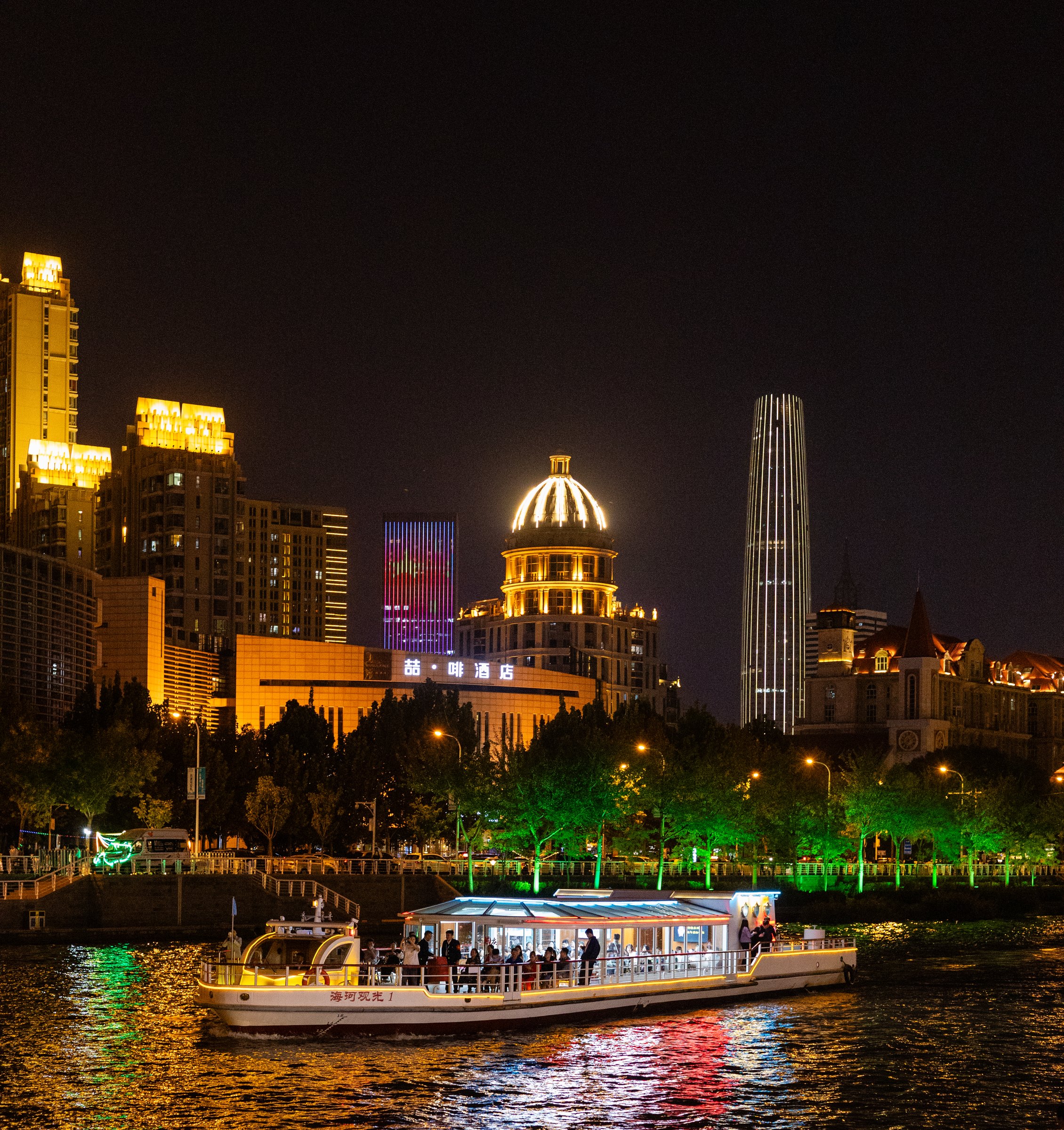 亮灯游船驶过夜间河面，岸边楼宇和树木映出金绿灯光